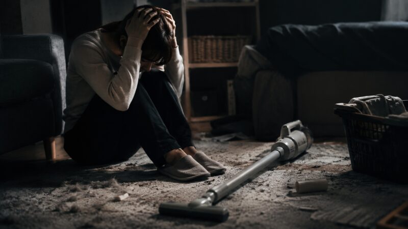 掃除機の前で悩み後悔する女性と、散らかった部屋に置かれたコードレス掃除機