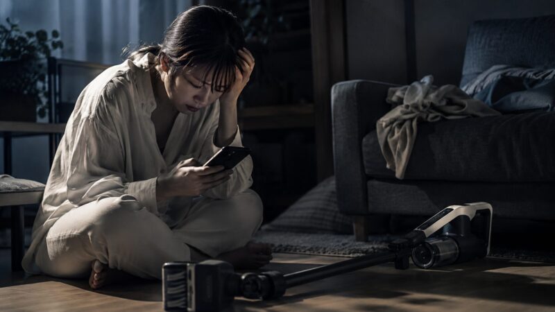 ラクかるスティック PV-BL3Mの口コミや評判をスマホで調べて後悔や不安を感じる女性と、リビングに置かれた一般的なデザインのコードレス掃除機を描いたシリアスなイメージ