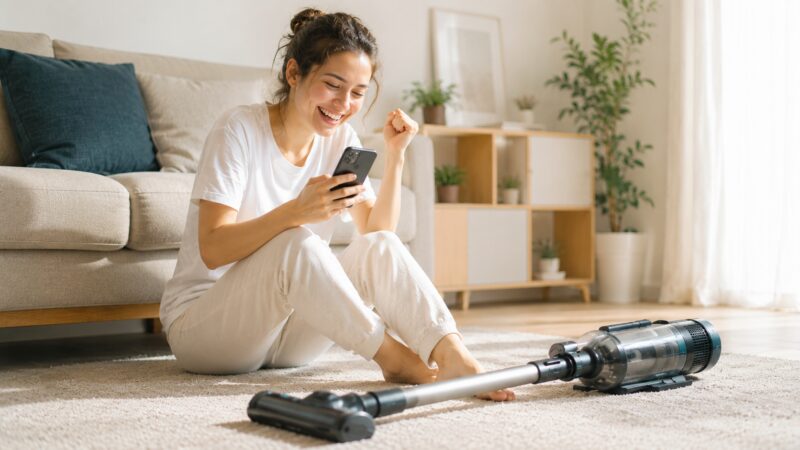 コードレス掃除機の口コミをスマホで確認して笑顔で喜ぶ女性、明るいリビングで掃除機を前に満足している様子