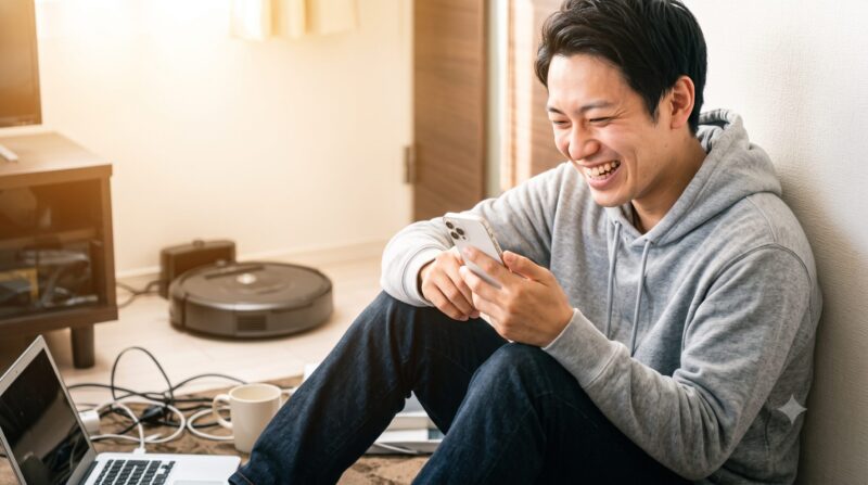 スマートフォンを操作しながら、思いっきり笑顔で笑っている若い日本人男性。リビングルーム、パーカー姿。