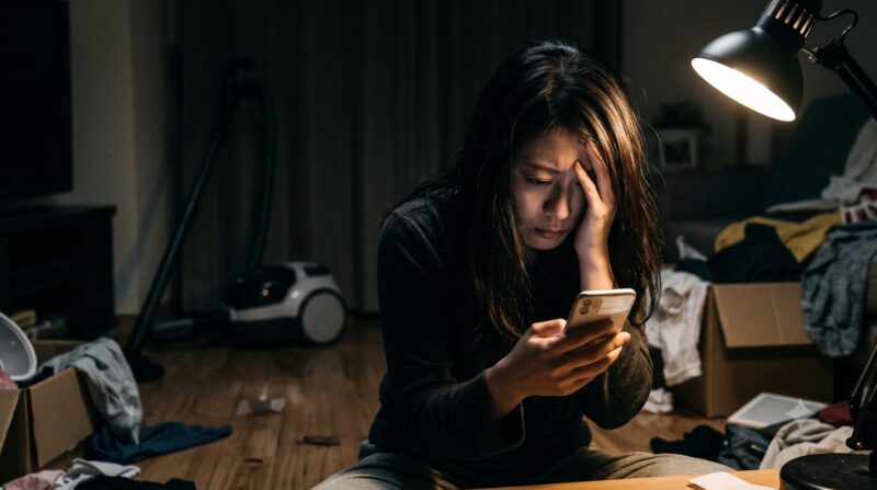 散らかった部屋でキャニスター掃除機の口コミをスマホで見て絶望し、悩む女性。