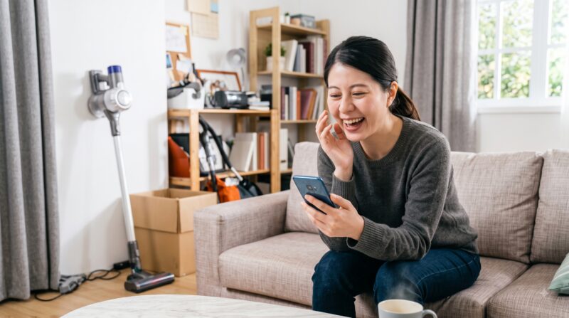明るいリビングで、ソファに座りスマートフォンを見て楽しそうに笑う女性。背景にコードレス掃除機。