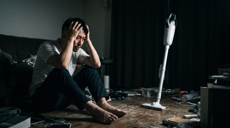 散らかった部屋で、一般的なコードレス掃除機を前に頭を抱えて絶望する日本人男性のドラマチックな写真