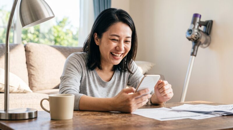 明るいリビングルームで、スマートフォンを手に満面の笑みを浮かべる日本人女性の様子。背景にはコードレス掃除機が見える。